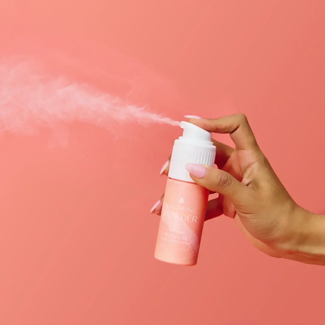 Mujer aplicando polvo voluminizador en cabello con fondo rosa, resaltando su textura y volumen natural.